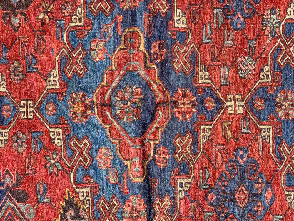 An antique Caucasian Soumak wool rug featuring geometric stepped medallions on a terracotta red field with blue and multi-coloured borders. View VI