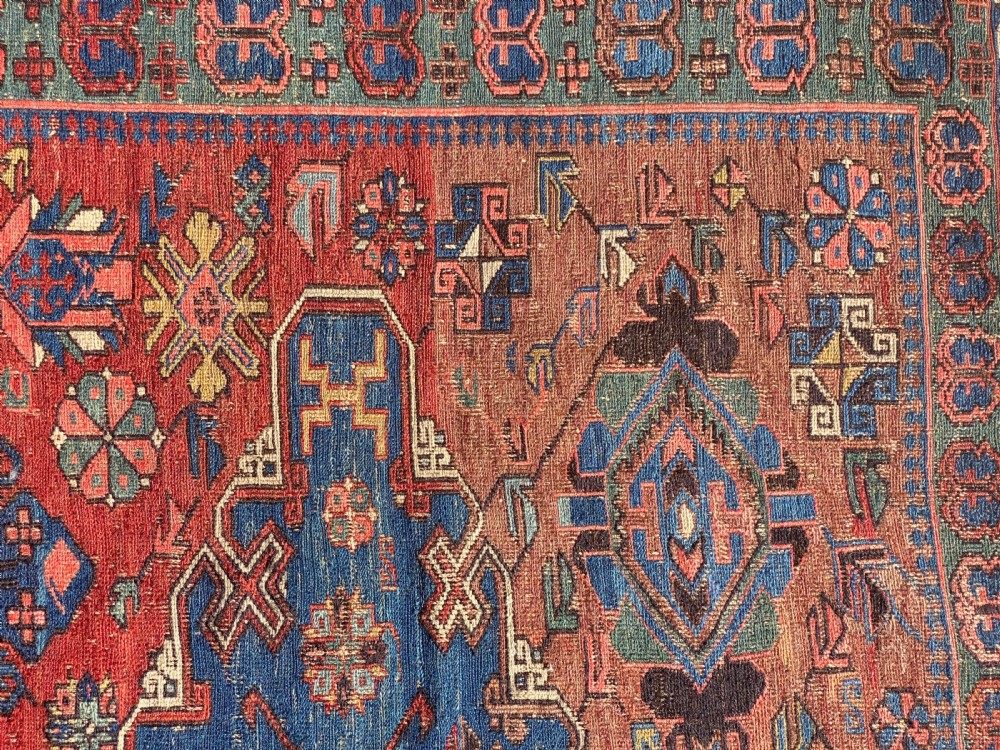 An antique Caucasian Soumak wool rug featuring geometric stepped medallions on a terracotta red field with blue and multi-coloured borders. View VII