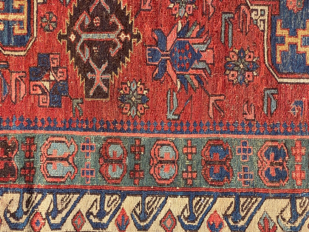 An antique Caucasian Soumak wool rug featuring geometric stepped medallions on a terracotta red field with blue and multi-coloured borders. View IX