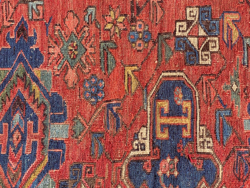 An antique Caucasian Soumak wool rug featuring geometric stepped medallions on a terracotta red field with blue and multi-coloured borders. View X