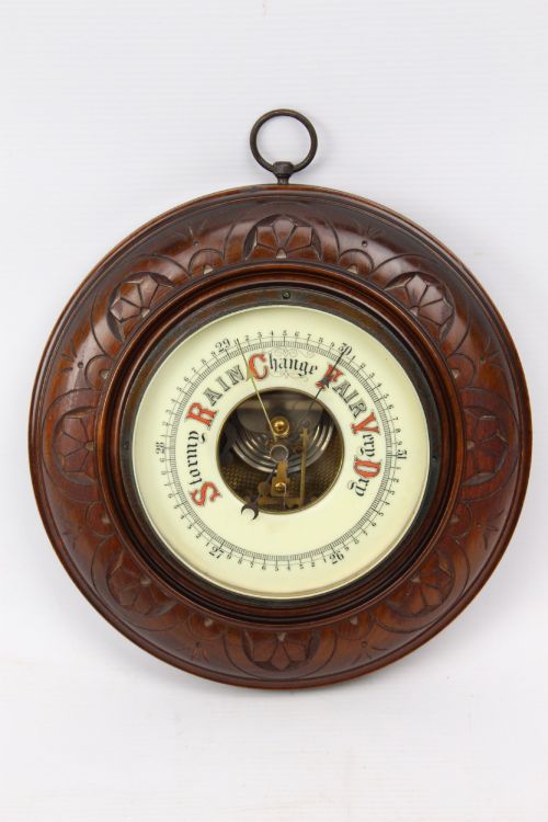 fig 1. A circular Victorian walnut wall barometer featuring a carved wooden border, a brass hanging ring, and an exposed brass skeleton movement within an enamel dial.