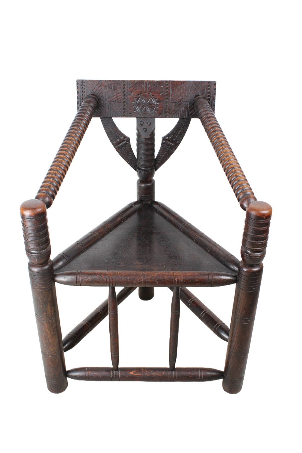 An antique dark brown oak turners chair featuring a triangular seat, bobbin-turned legs and arms, and a carved geometric back crest panel.