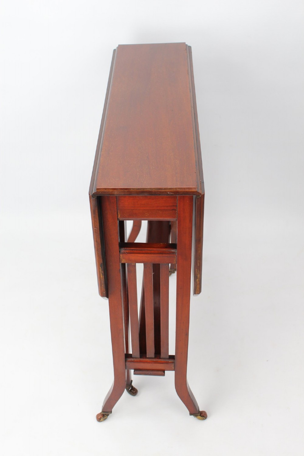 A compact Edwardian mahogany Sutherland table with folded side leaves, slatted end supports, and legs fitted with brass castors. View II