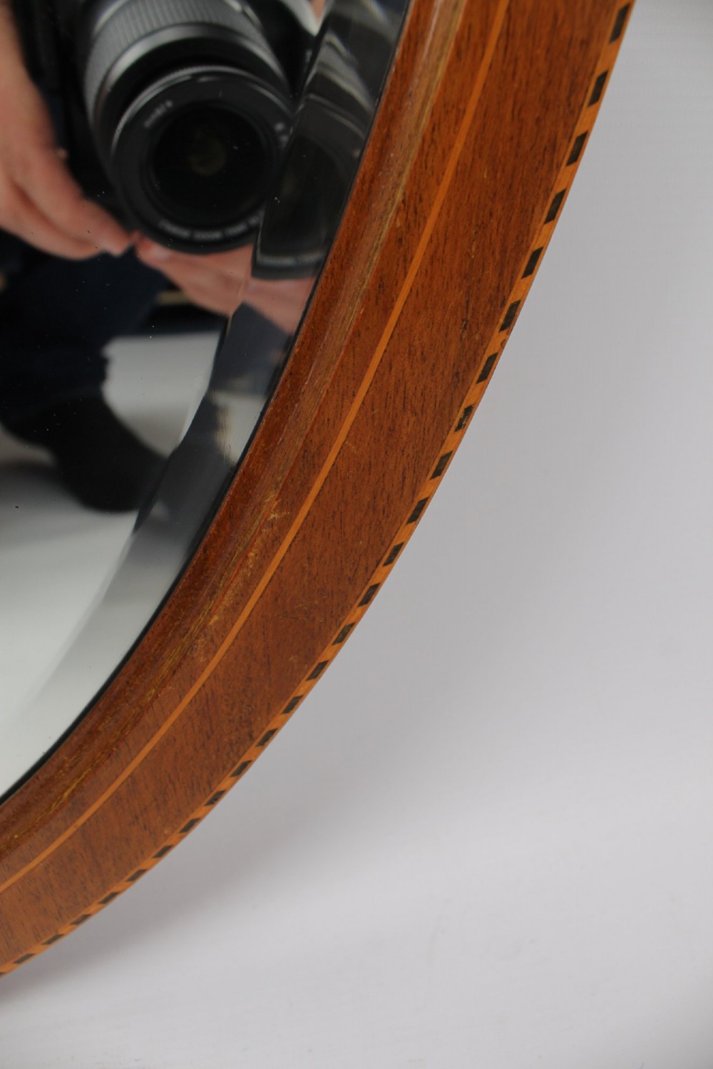 An oval wall mirror framed in warm mahogany timber with geometric chequered inlay along the outer border. View IV