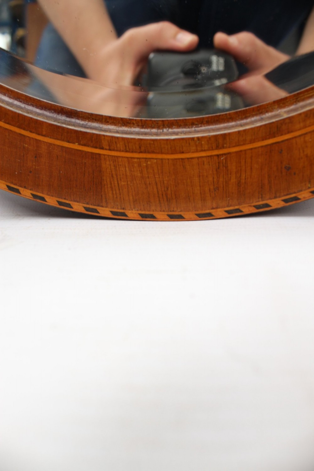 An oval wall mirror framed in warm mahogany timber with geometric chequered inlay along the outer border. View VI