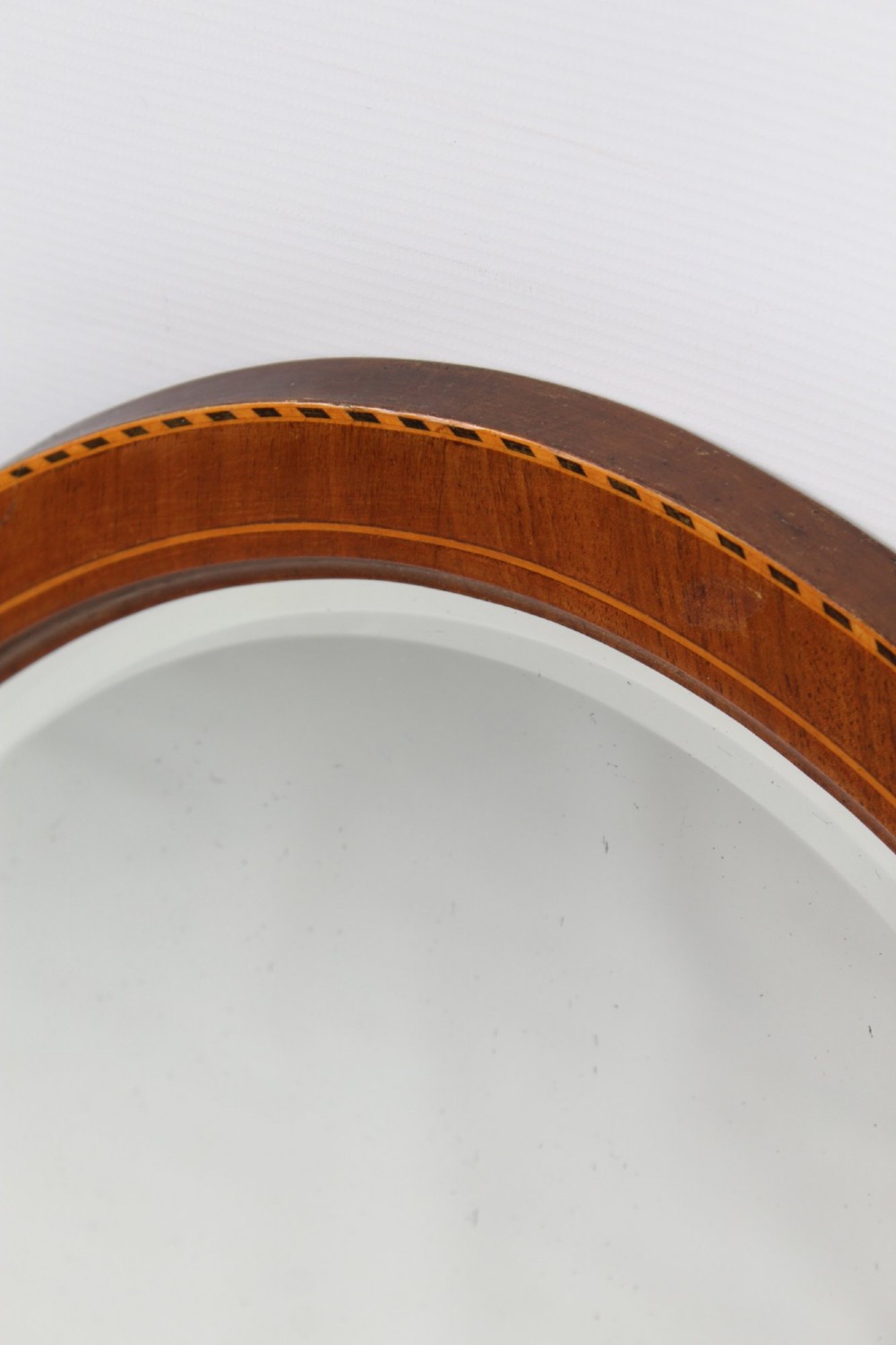An oval wall mirror framed in warm mahogany timber with geometric chequered inlay along the outer border. View VII
