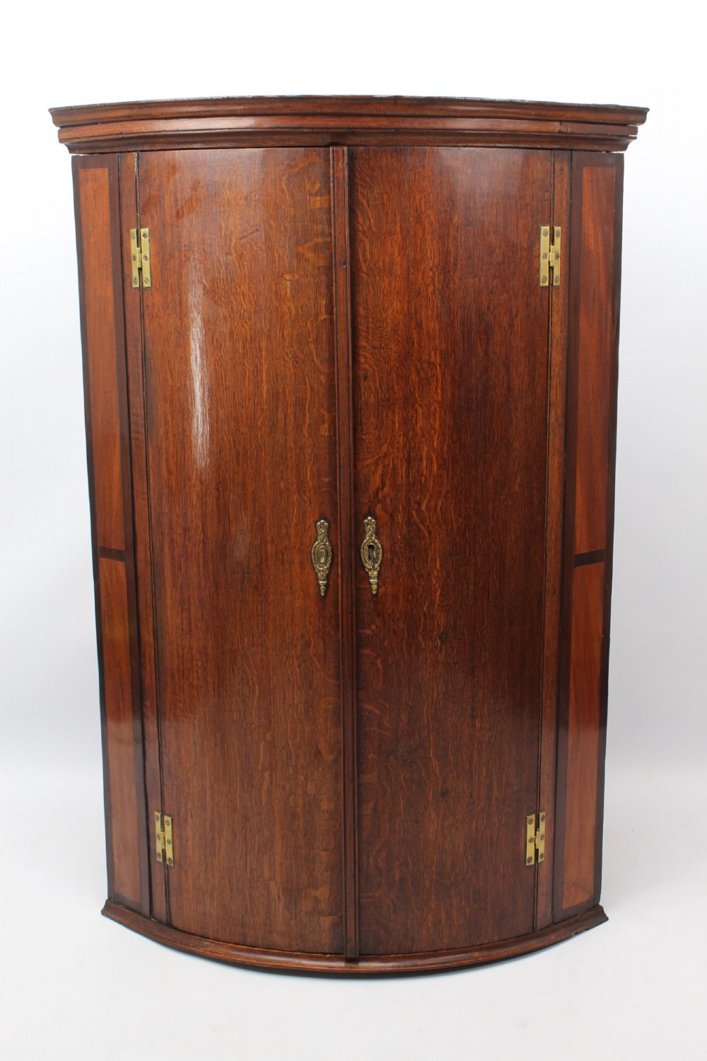 A Georgian oak bow-front corner cupboard with twin doors, exposed brass H-hinges, crossbanded veneer borders, and central brass escutcheons.