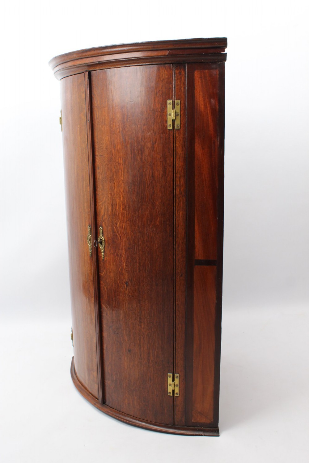 A Georgian oak bow-front corner cupboard with twin doors, exposed brass H-hinges, crossbanded veneer borders, and central brass escutcheons. View II
