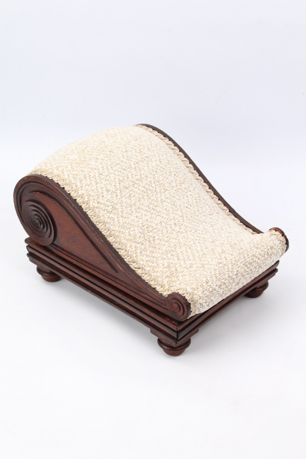 An inclined Regency rosewood footstool featuring carved side scrolls, turned bun feet, and cream textured upholstery finished with gimp braid.