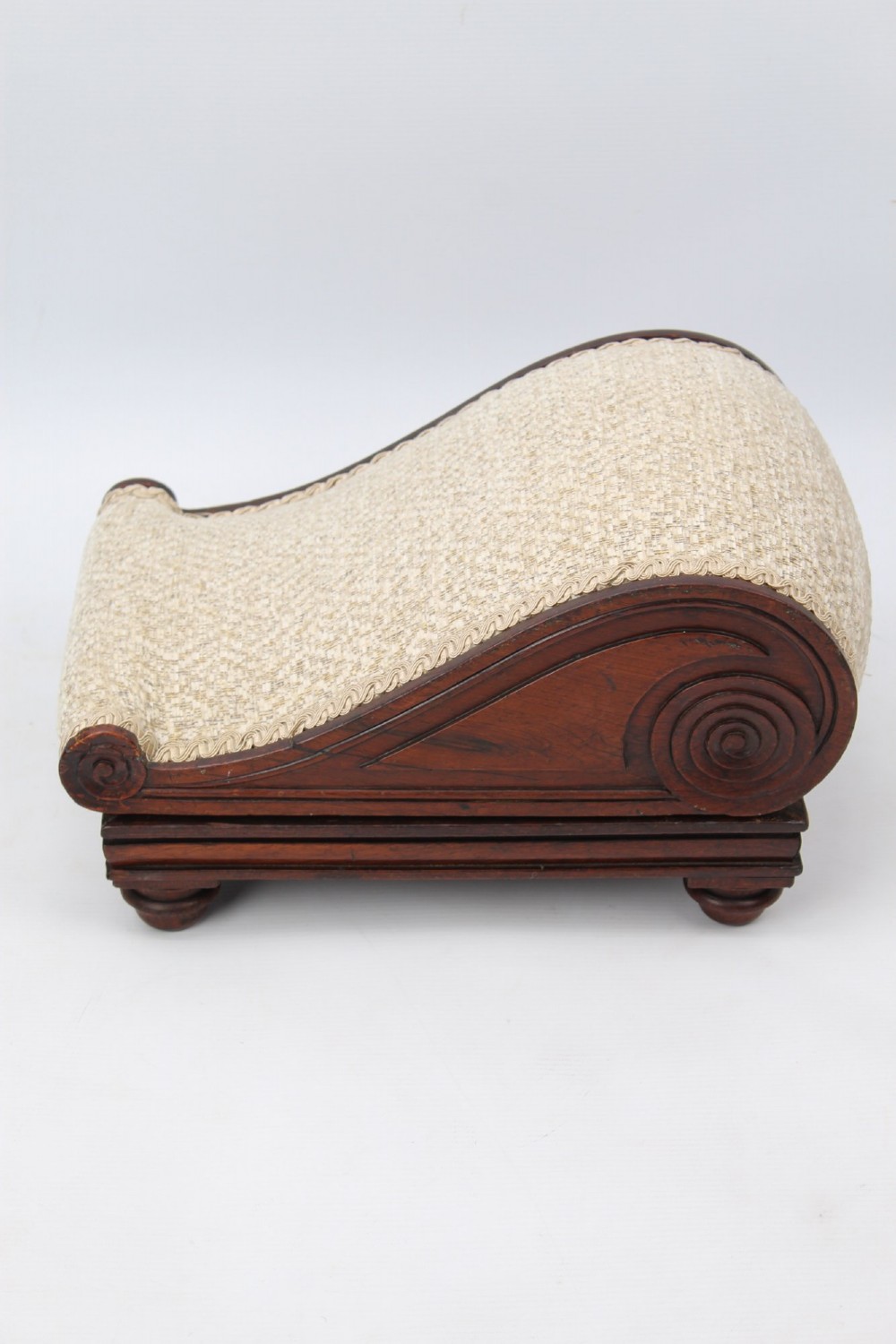 An inclined Regency rosewood footstool featuring carved side scrolls, turned bun feet, and cream textured upholstery finished with gimp braid. View IV