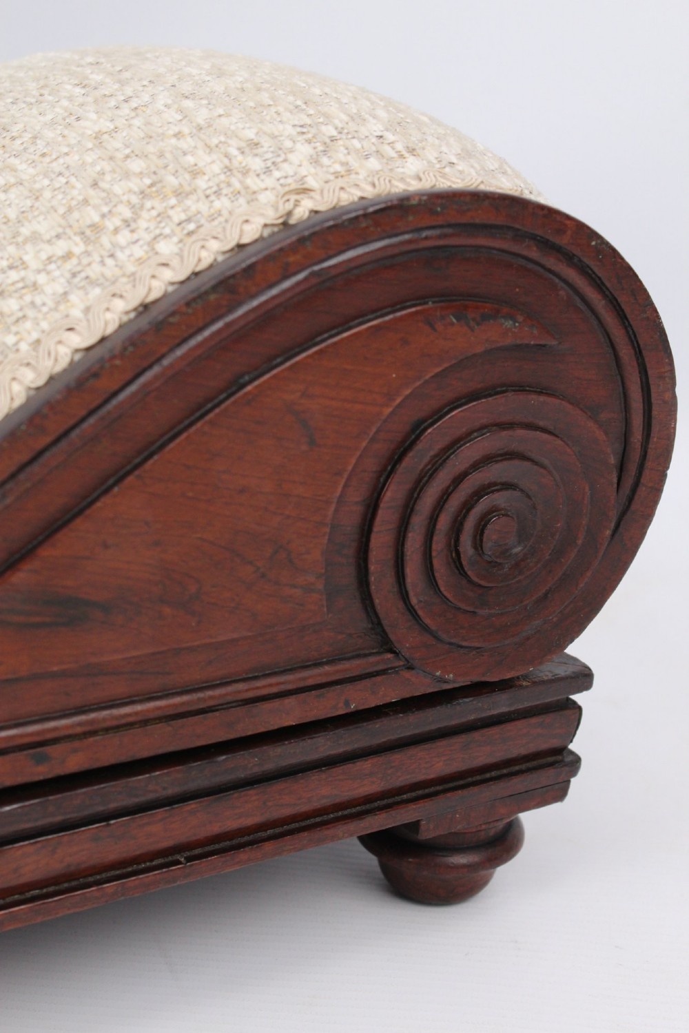 An inclined Regency rosewood footstool featuring carved side scrolls, turned bun feet, and cream textured upholstery finished with gimp braid. View VII