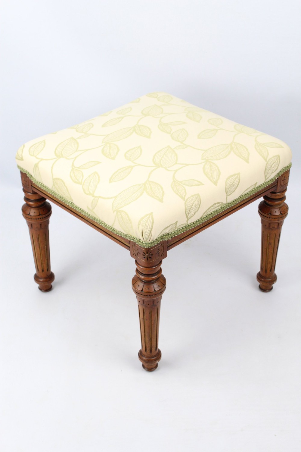 A square Victorian oak dressing stool featuring fluted turned legs with gilt accents and a cream fabric seat decorated with green leaf motifs.