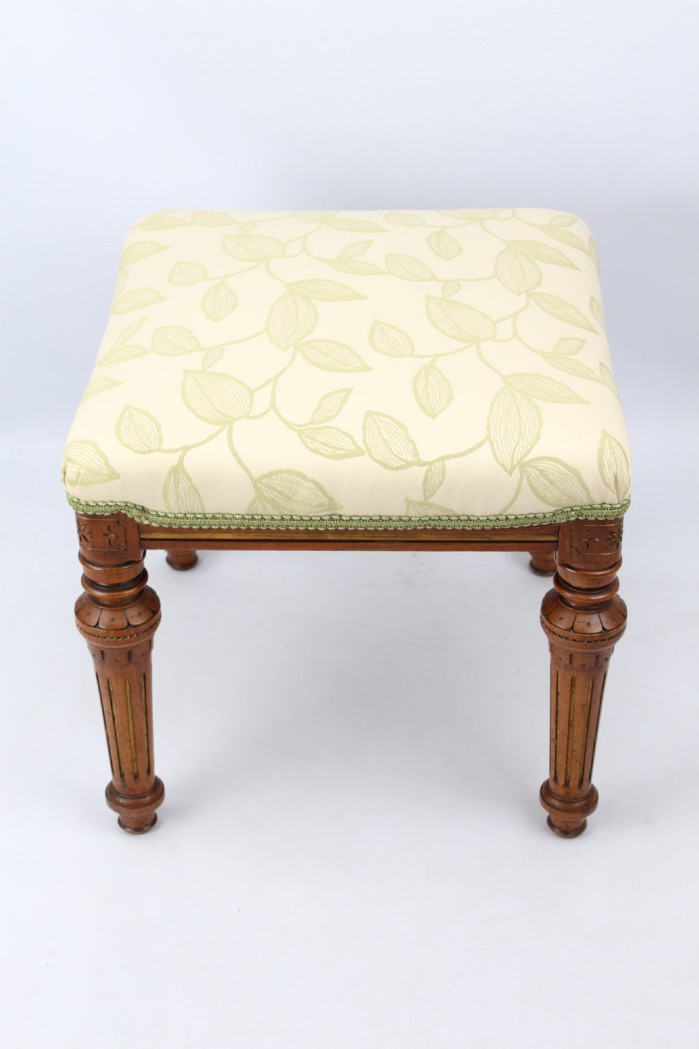 A square Victorian oak dressing stool featuring fluted turned legs with gilt accents and a cream fabric seat decorated with green leaf motifs. View II