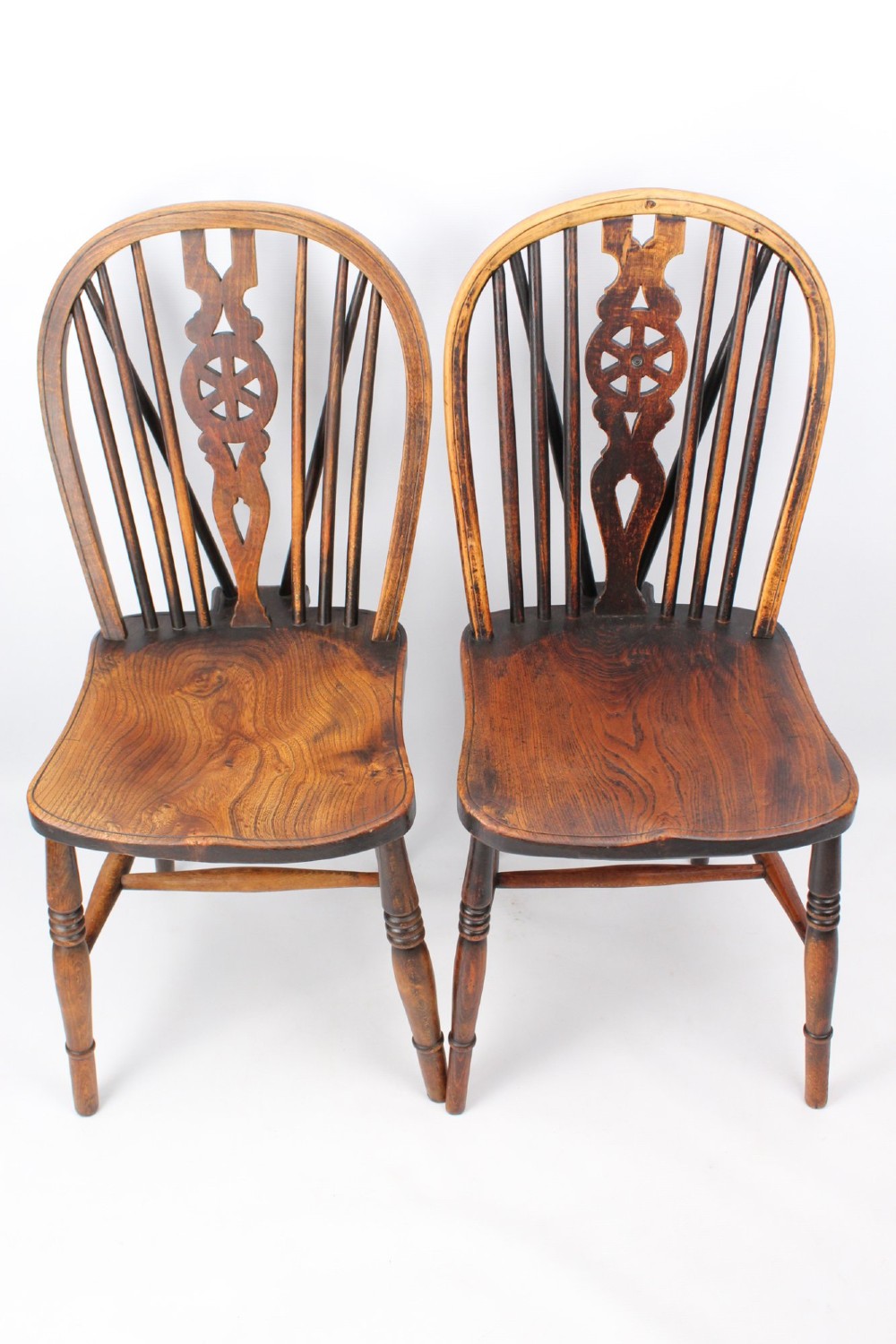 A near pair of antique dark timber Windsor kitchen chairs featuring pierced wheelback splats, contoured saddle seats, and turned legs.