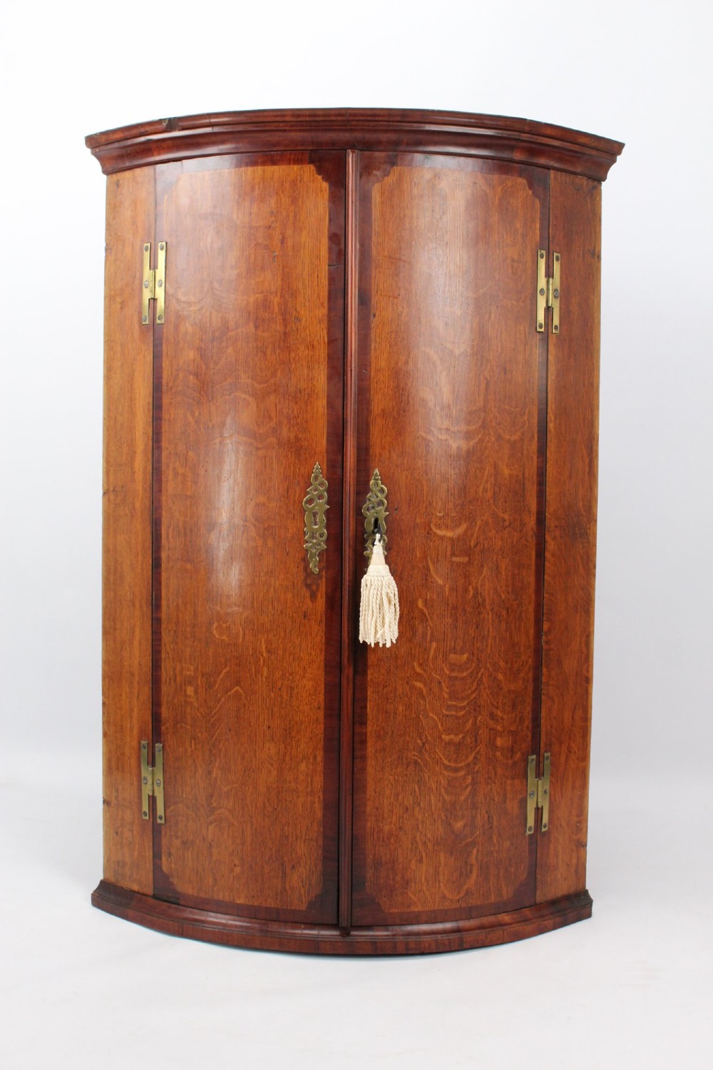 An 18th-century Georgian oak corner cupboard featuring a curved bow front, brass H-hinges, keyhole escutcheon plates, and mahogany crossbanding.