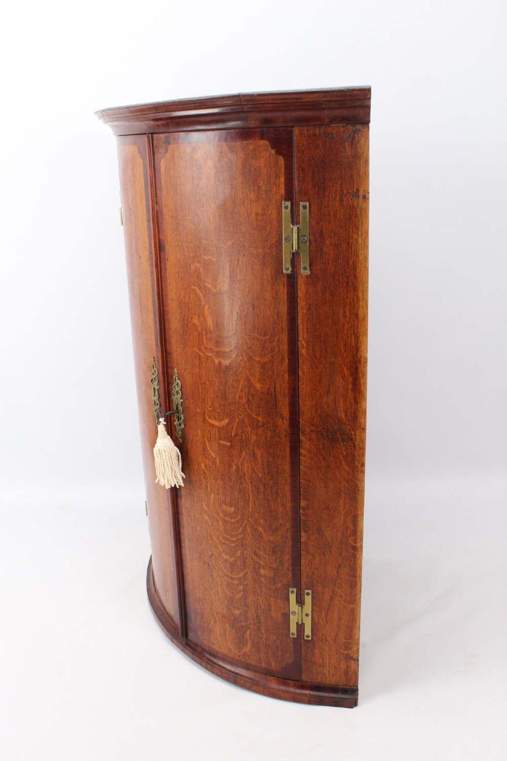 An 18th-century Georgian oak corner cupboard featuring a curved bow front, brass H-hinges, keyhole escutcheon plates, and mahogany crossbanding. View II