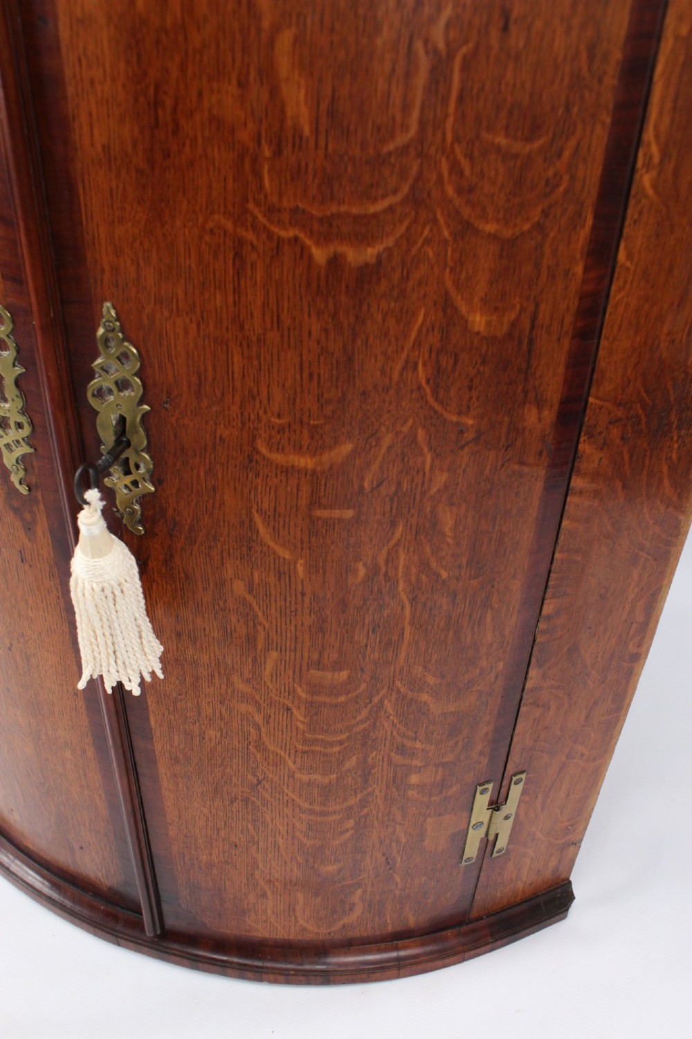 An 18th-century Georgian oak corner cupboard featuring a curved bow front, brass H-hinges, keyhole escutcheon plates, and mahogany crossbanding. View VII