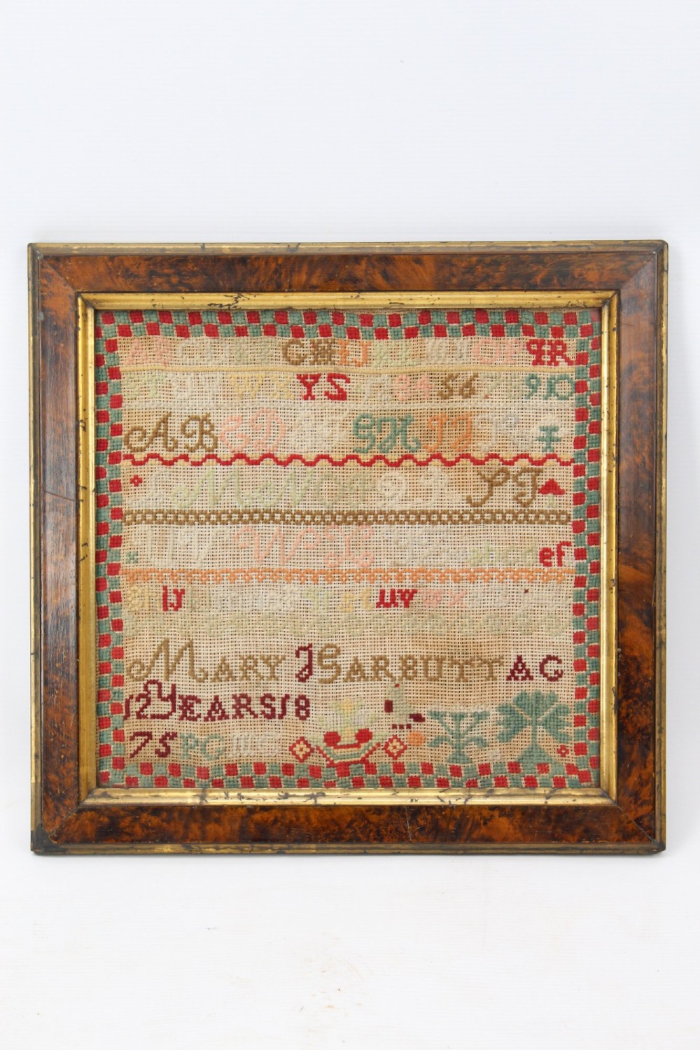 A framed 1875 Victorian needlework sampler featuring rows of embroidered alphabets, geometric borders, and text reading Mary J Garbutt Aged 12 Years inside a wooden frame.