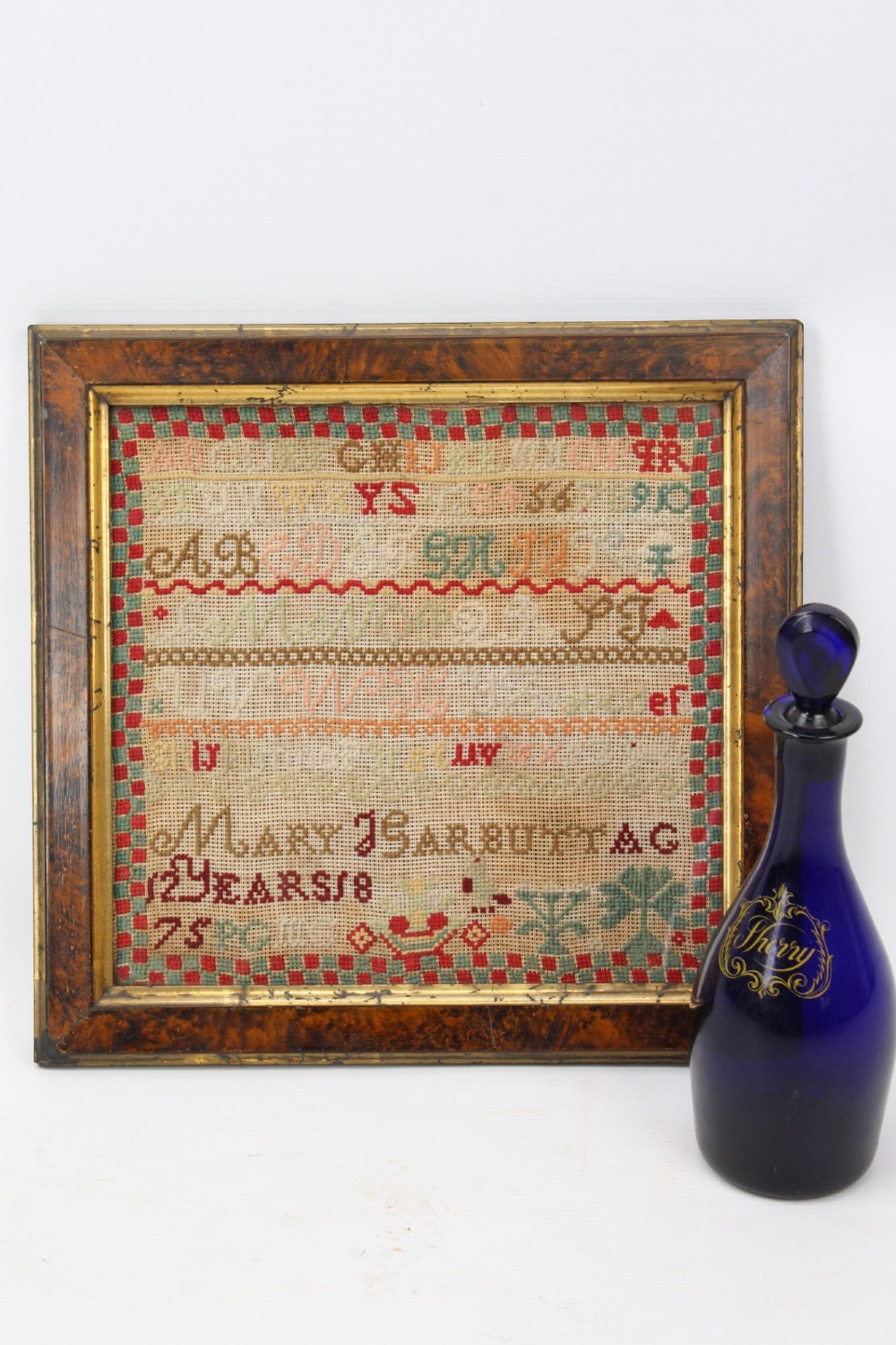 A framed 1875 Victorian needlework sampler featuring rows of embroidered alphabets, geometric borders, and text reading Mary J Garbutt Aged 12 Years inside a wooden frame. View II