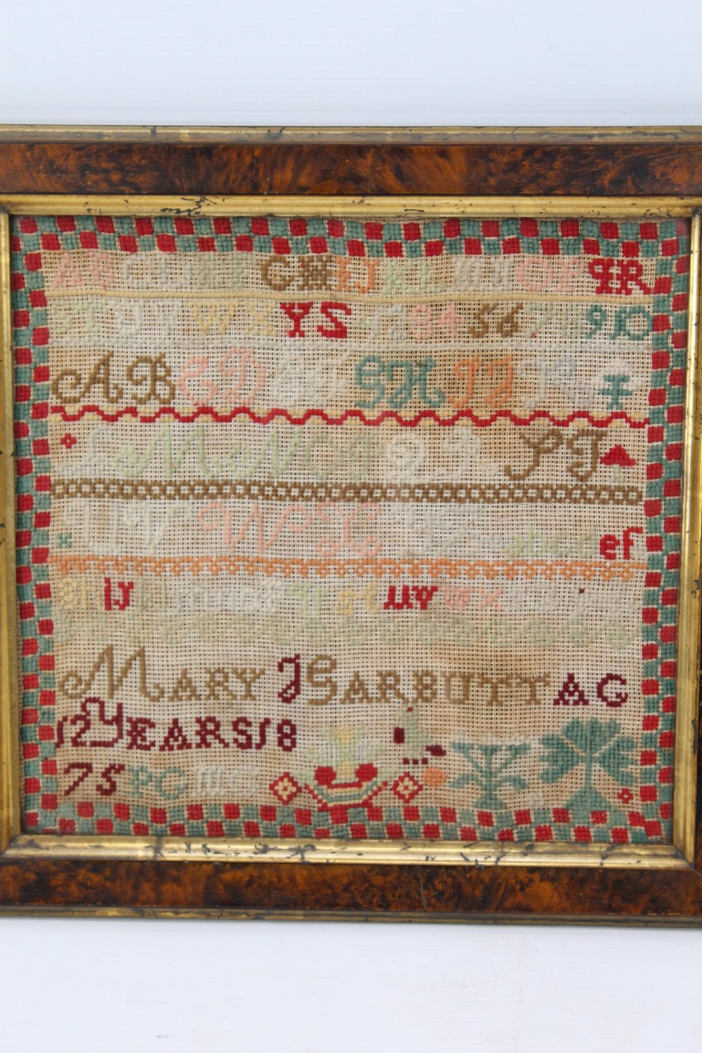 A framed 1875 Victorian needlework sampler featuring rows of embroidered alphabets, geometric borders, and text reading Mary J Garbutt Aged 12 Years inside a wooden frame. View III
