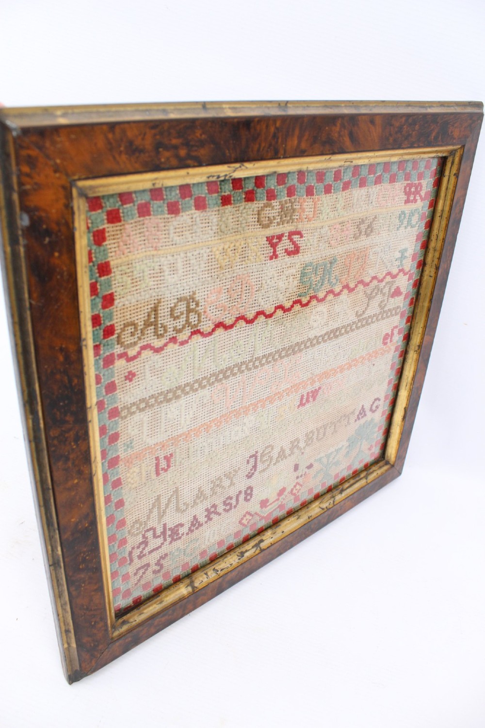 A framed 1875 Victorian needlework sampler featuring rows of embroidered alphabets, geometric borders, and text reading Mary J Garbutt Aged 12 Years inside a wooden frame. View IV