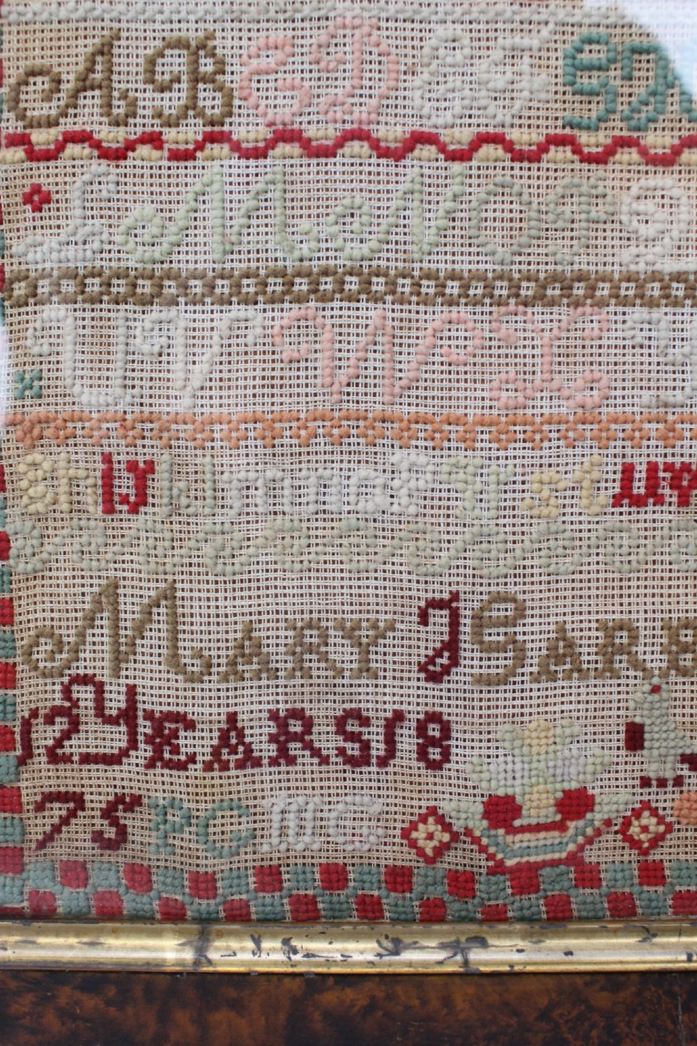 A framed 1875 Victorian needlework sampler featuring rows of embroidered alphabets, geometric borders, and text reading Mary J Garbutt Aged 12 Years inside a wooden frame. View X