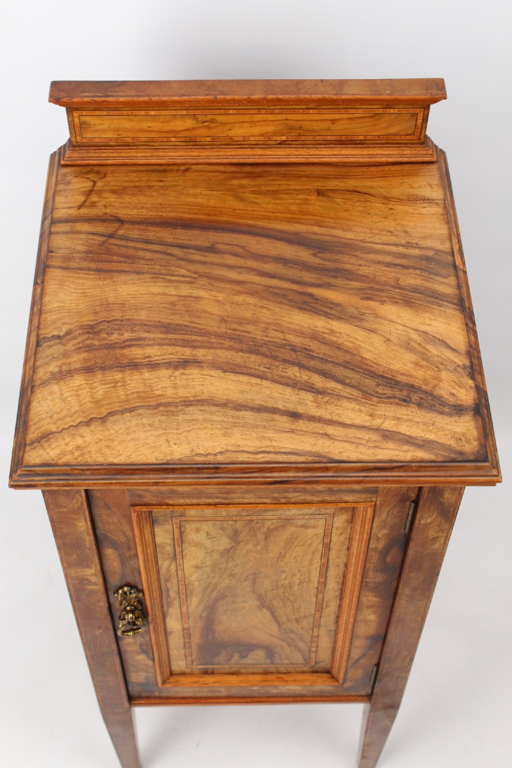 An Edwardian burr walnut bedside cabinet featuring a small gallery top, crossbanded inlaid door panel, drop brass handle, and square tapered legs. View VI