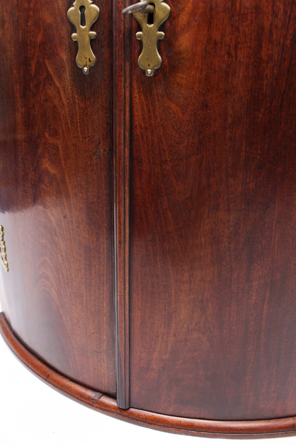 A Georgian mahogany bow front corner cupboard with twin curved doors and decorative brass hinges and keyhole escutcheons. View XII