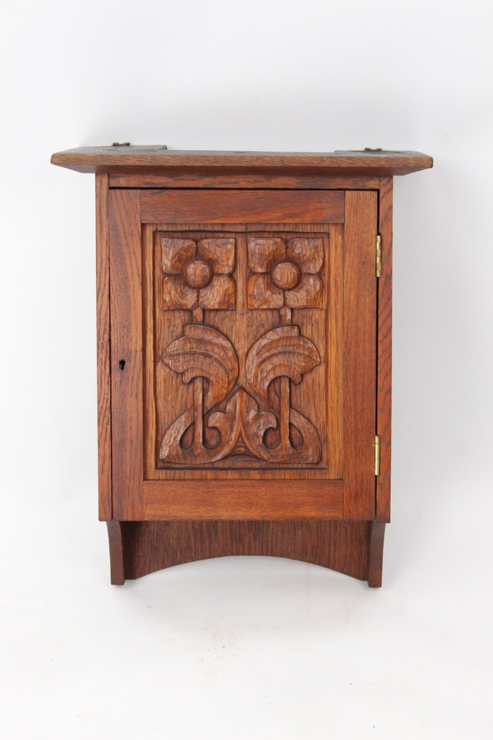 A small Edwardian Arts and Crafts oak wall cabinet featuring a single door decorated with a carved stylized floral panel and small brass hinges.