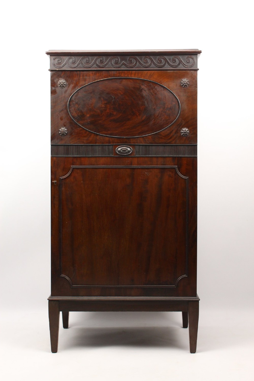 An Edwardian dark mahogany pedestal cabinet featuring an oval flame mahogany panel, carved scroll frieze, and square tapered legs.