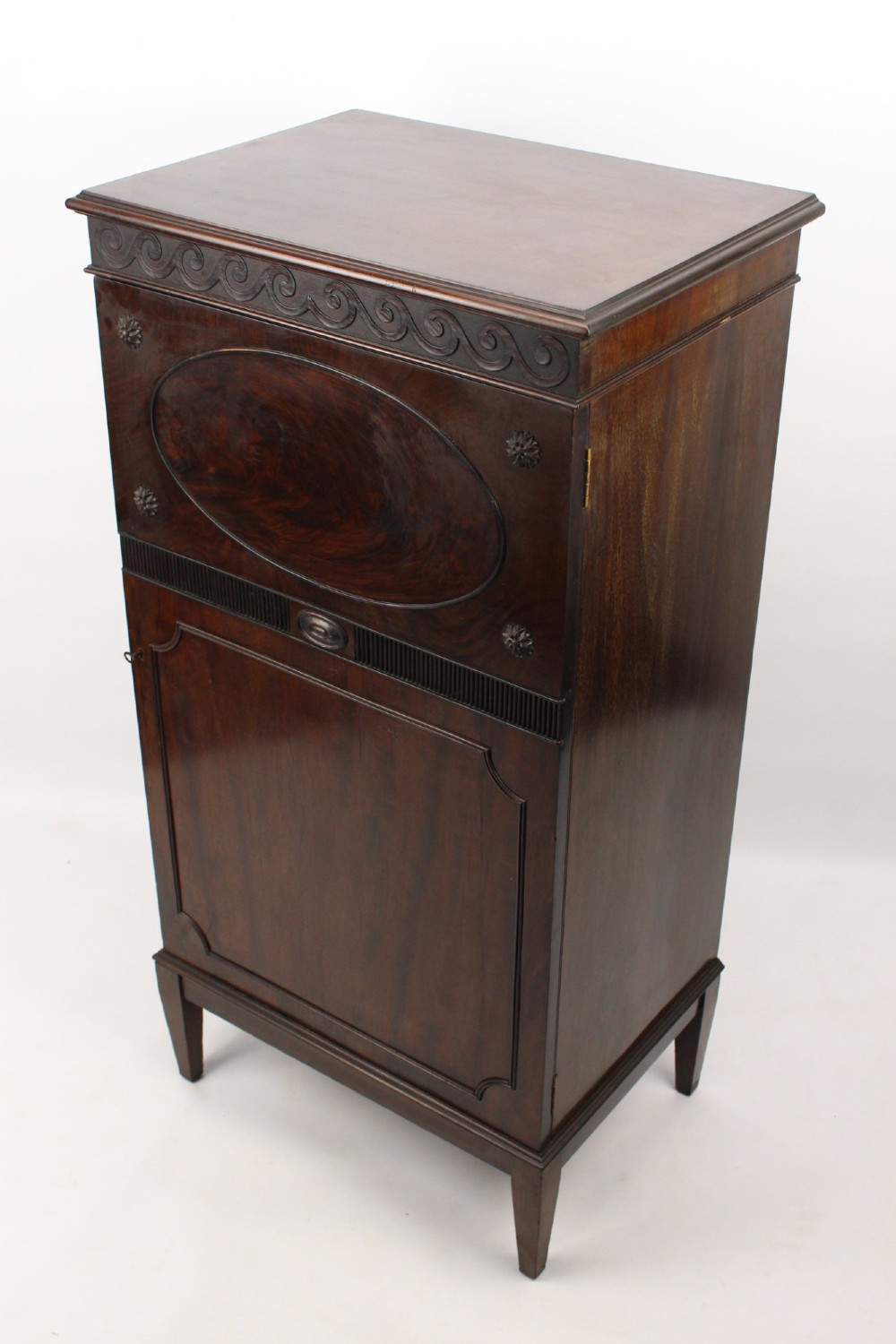An Edwardian dark mahogany pedestal cabinet featuring an oval flame mahogany panel, carved scroll frieze, and square tapered legs. View IV