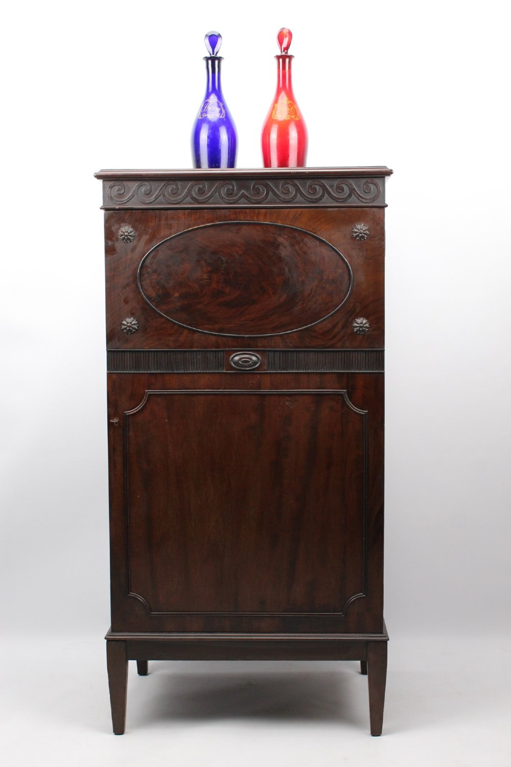 An Edwardian dark mahogany pedestal cabinet featuring an oval flame mahogany panel, carved scroll frieze, and square tapered legs. View VII