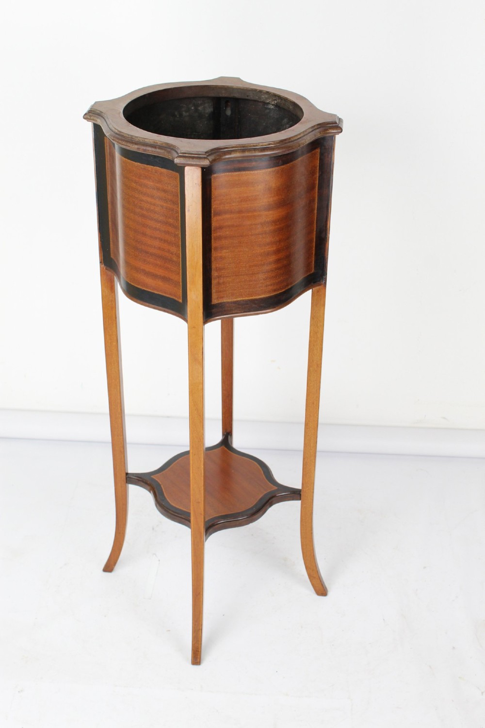 A tall Edwardian mahogany plant stand featuring shaped inlaid panels, four slender splayed legs, a lower shelf, and a dark metal liner.