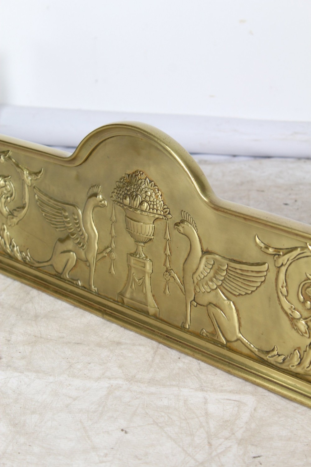A long Victorian brass fireplace fender featuring embossed Neoclassical relief decoration of central urns, winged sphinxes, and foliate scrolls. View V