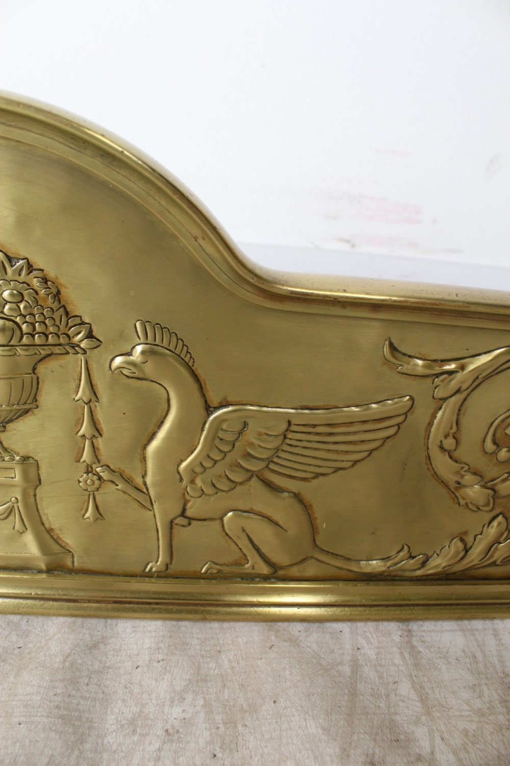 A long Victorian brass fireplace fender featuring embossed Neoclassical relief decoration of central urns, winged sphinxes, and foliate scrolls. View VI