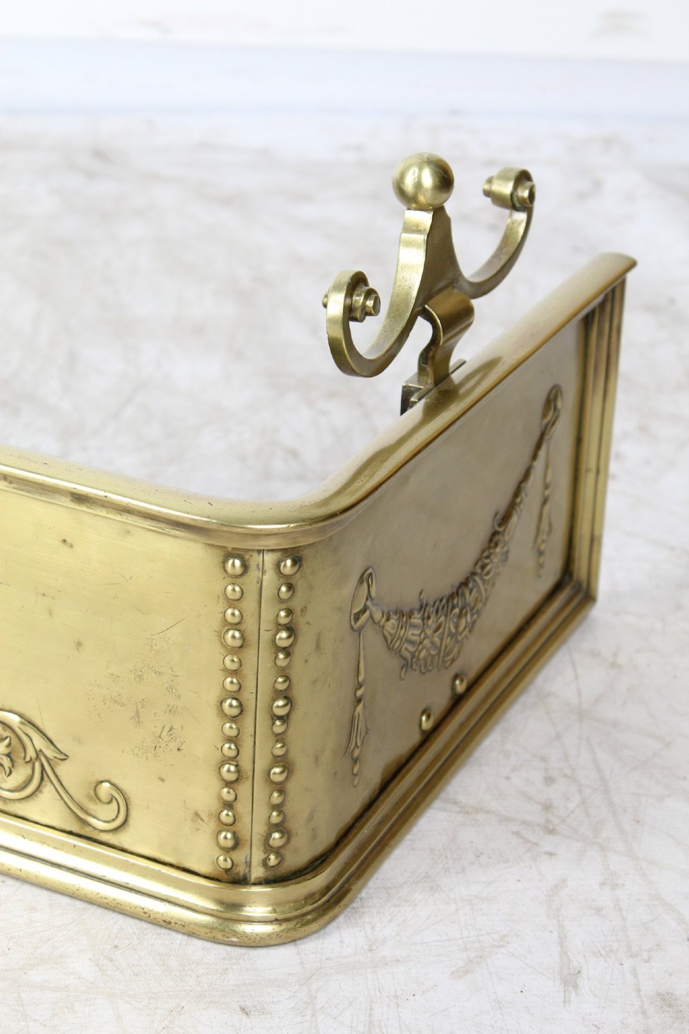 A long Victorian brass fireplace fender featuring embossed Neoclassical relief decoration of central urns, winged sphinxes, and foliate scrolls. View VII