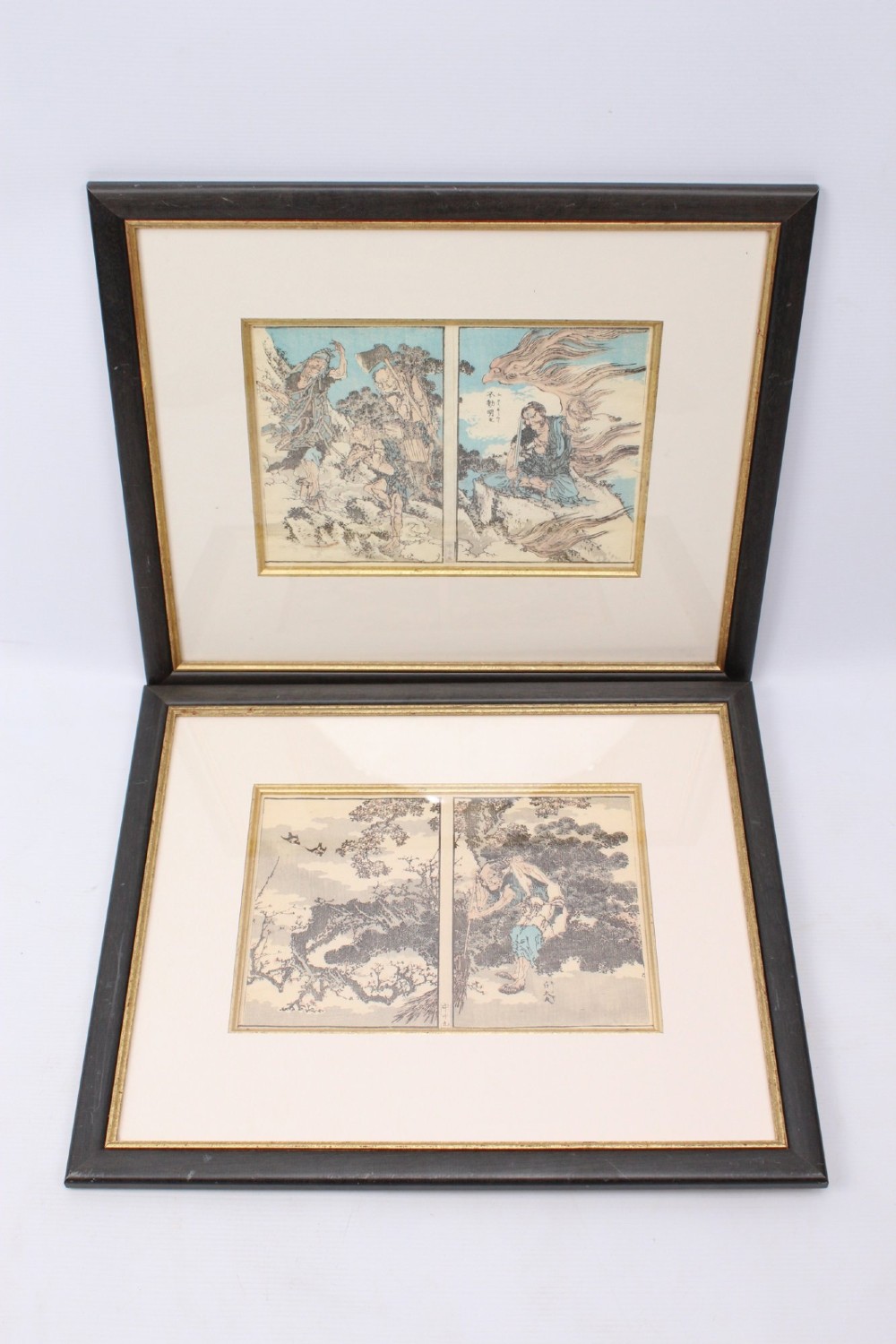 A pair of framed Japanese woodblock prints featuring detailed figurative and landscape illustrations mounted in cream mats within dark wooden frames.