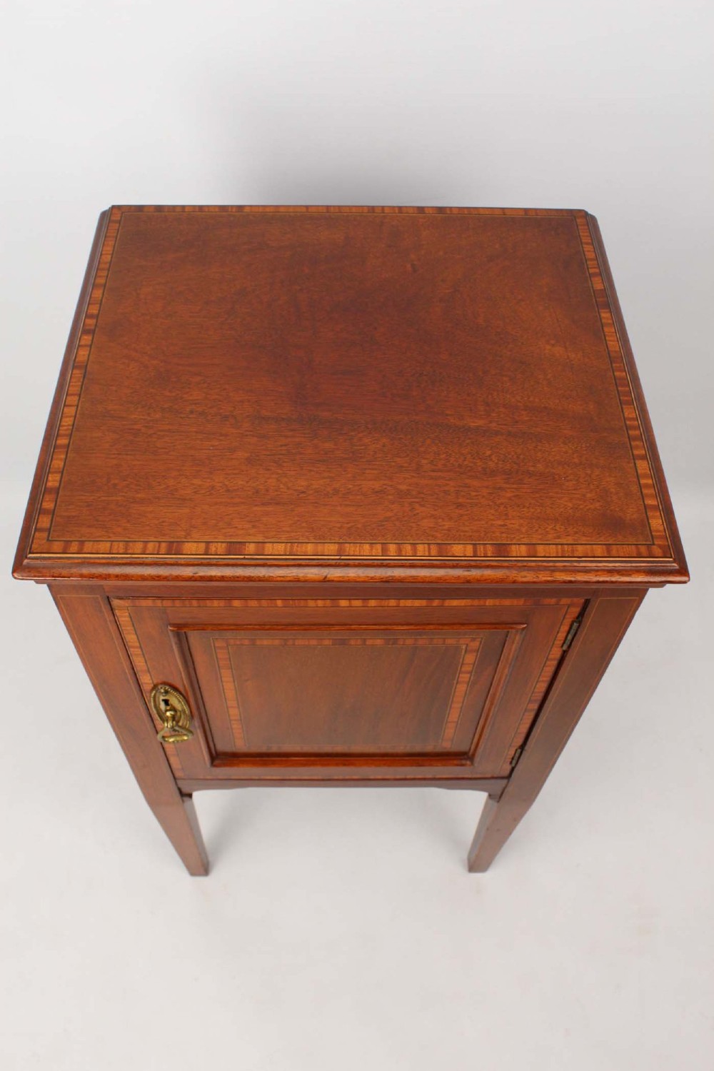 An Edwardian mahogany bedside cabinet featuring satinwood stringing inlay, a single panelled door with a brass handle, and square tapered legs. View VI