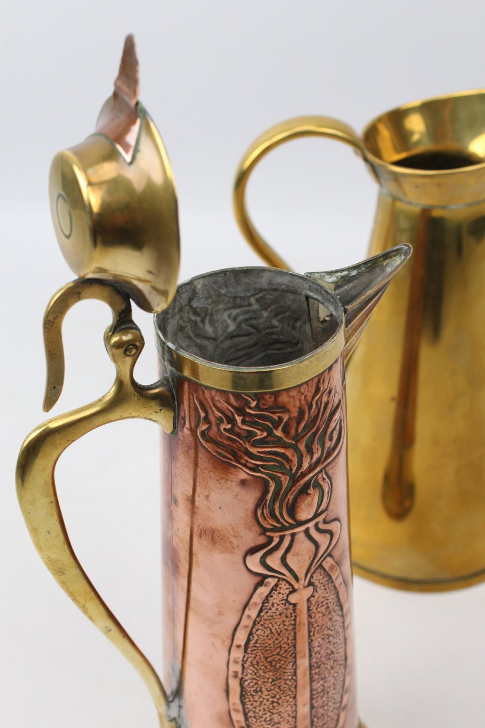 A pair of antique metal jugs, one crafted from repousse copper with brass fittings and a hinged lid alongside a tapered solid brass pitcher. View IX