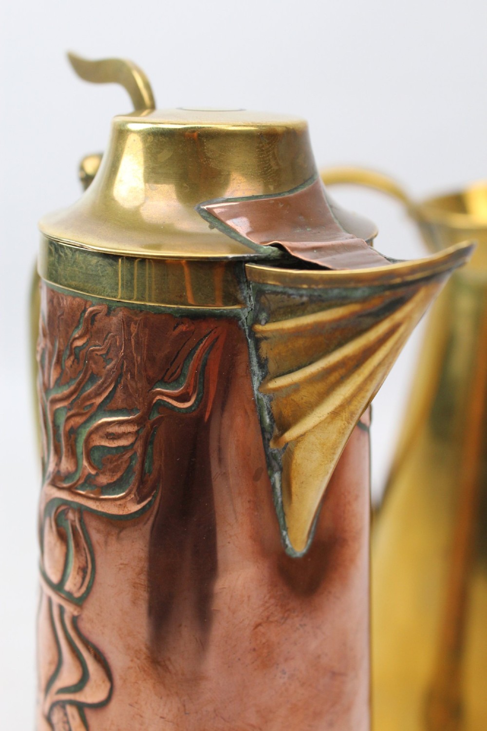 A pair of antique metal jugs, one crafted from repousse copper with brass fittings and a hinged lid alongside a tapered solid brass pitcher. View X