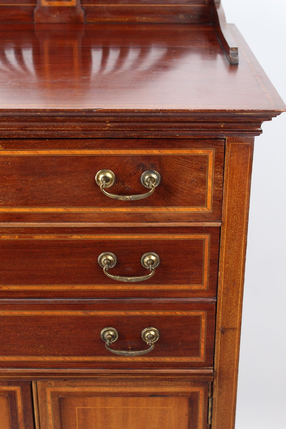 An Edwardian mahogany music cabinet featuring inlaid stringing, three top drawers with brass handles, a two-door lower cupboard, and a carved cresting gallery. View XII