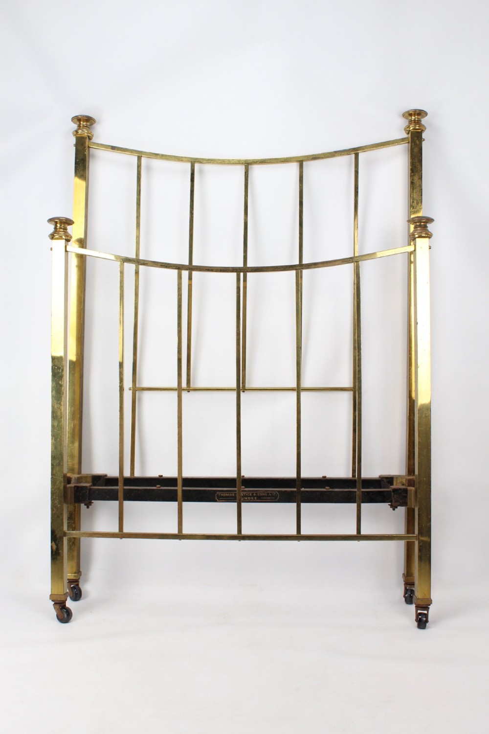 A Victorian brass single bed frame featuring a gently curved top rail, vertical spindles, turned finials, and original wheel castors.