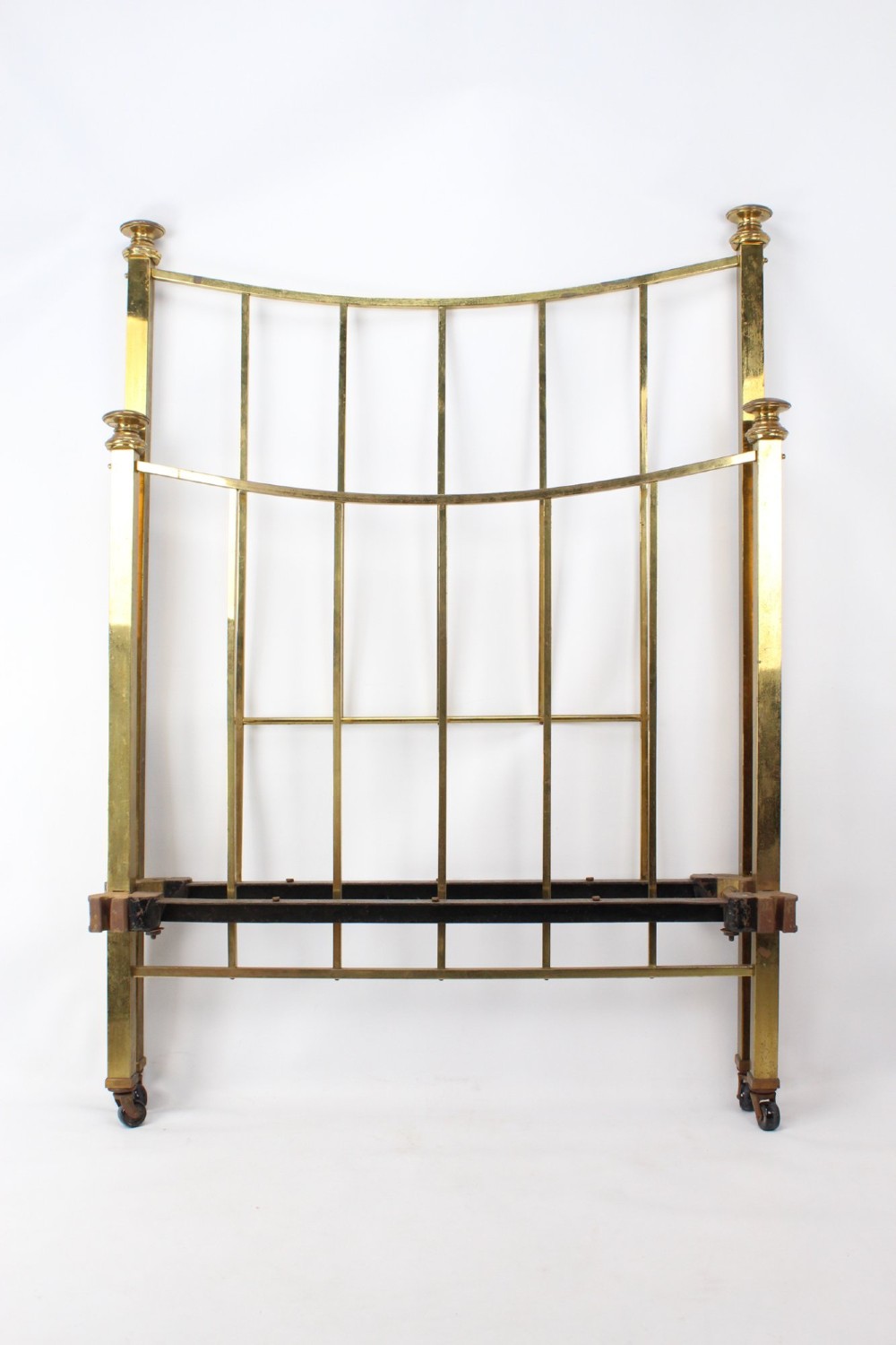 A Victorian brass single bed frame featuring a gently curved top rail, vertical spindles, turned finials, and original wheel castors. View II