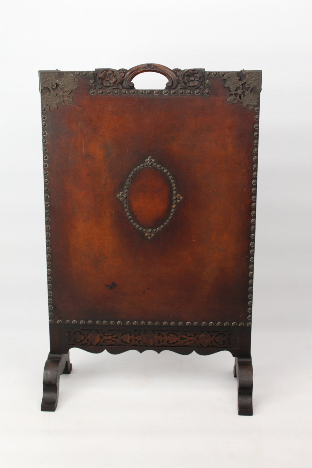 An Edwardian Arts and Crafts fire screen constructed from carved oak and warm brown leather featuring brass studwork and a central oval motif.