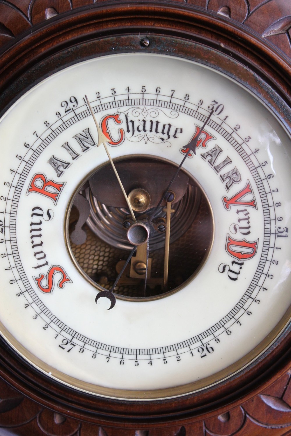 A circular Victorian walnut wall barometer featuring a carved wooden border, a brass hanging ring, and an exposed brass skeleton movement within an enamel dial. View IV