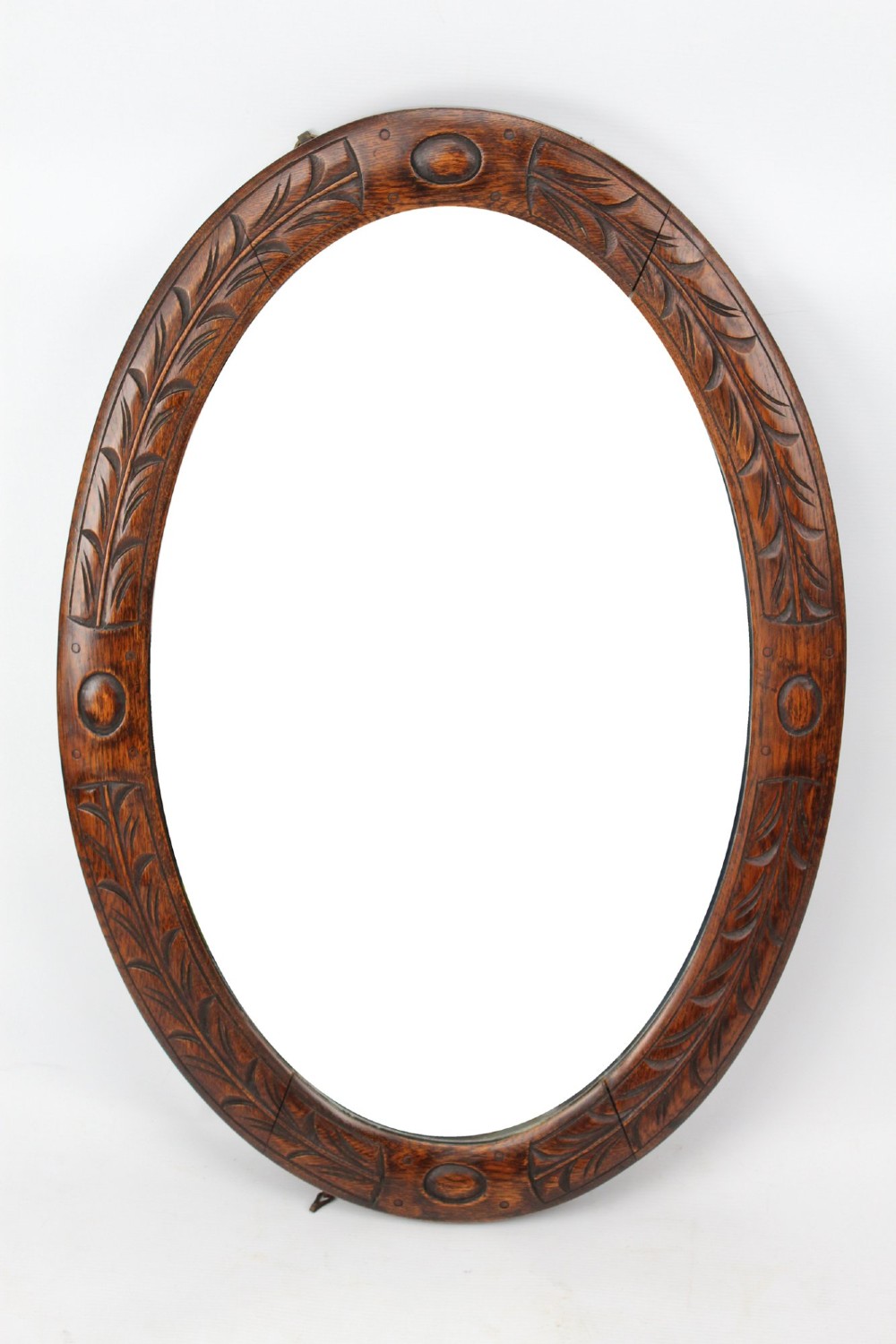 An Edwardian oval wall mirror featuring a carved dark oak frame decorated with leaf motifs and rounded bosses.