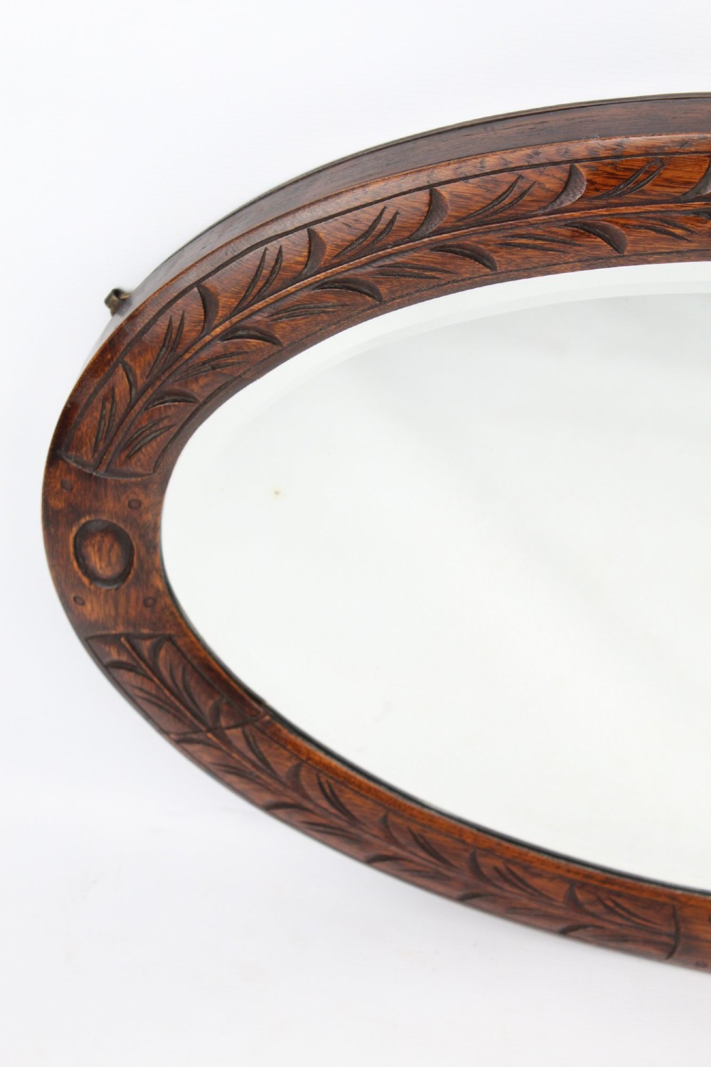 An Edwardian oval wall mirror featuring a carved dark oak frame decorated with leaf motifs and rounded bosses. View V