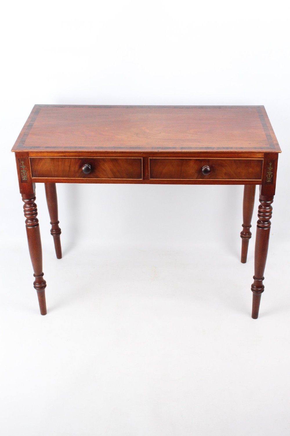 A Regency mahogany writing desk featuring brass inlaid corner stiles, two frieze drawers with wooden knobs, and turned legs.
