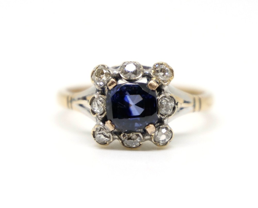 An 18ct gold ring featuring a central cushion-cut blue sapphire framed by eight round diamonds in a white metal cluster setting.