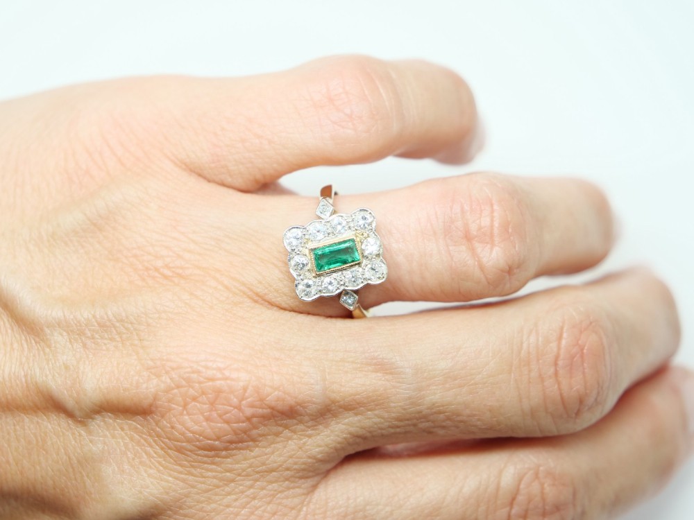 An Art Deco style ring featuring a central emerald-cut green emerald surrounded by a scalloped border of round diamonds set in white metal on a yellow gold band. View V